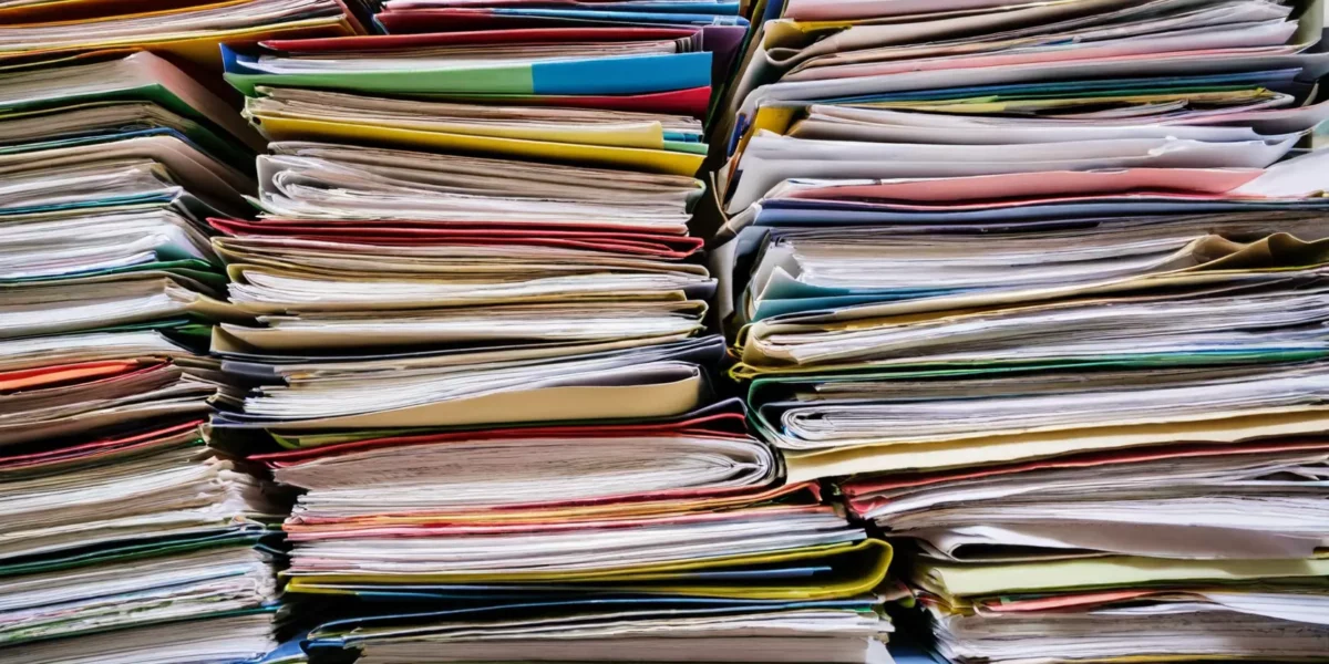 Pile ordinate di fascicoli e documenti multicolori impilati in colonne fitte, con fogli sovrapposti e copertine piegate in un archivio caotico.