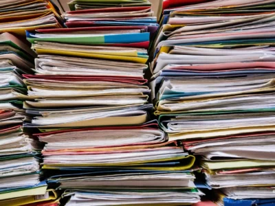 Pile ordinate di fascicoli e documenti multicolori impilati in colonne fitte, con fogli sovrapposti e copertine piegate in un archivio caotico.